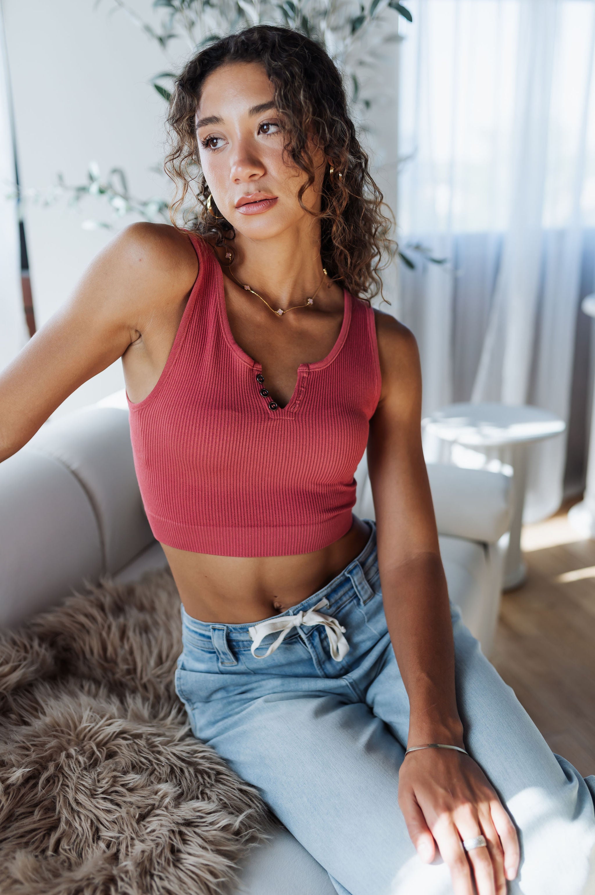 Woman wearing a red crop top and blue jeans sitting on a couch in a bright room.