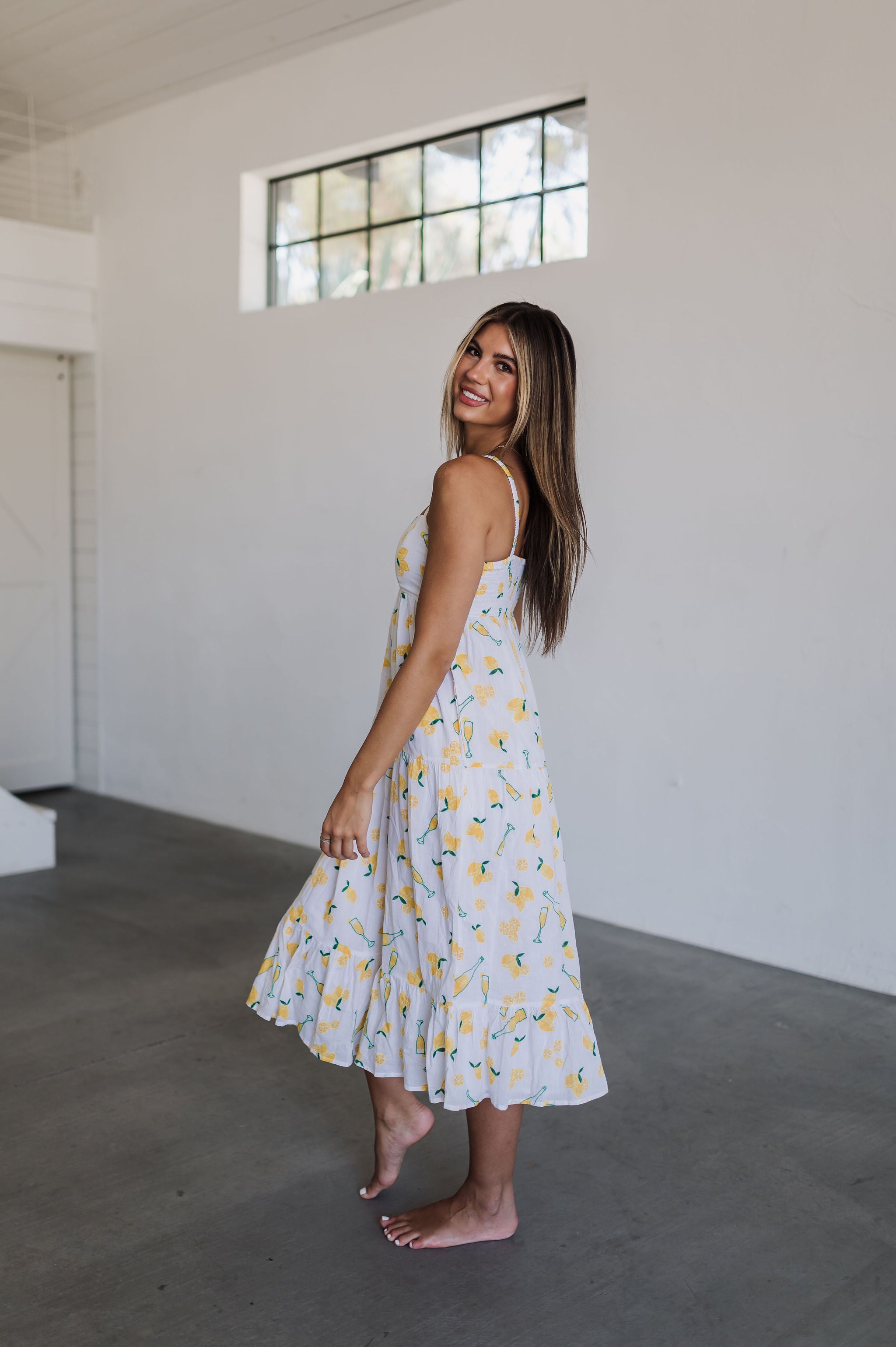 Side view of Summer Fling Midi Dress with embroidered Limoncello print, thin straps, square neckline, side pockets, and ruffled hem.