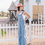 Side angled view of Clementine Wide Leg Distressed Denim Overalls with raw hem and front and side pockets. 
