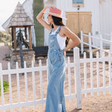 Side angled view of Clementine Wide Leg Distressed Denim Overalls with raw hem and front and side pockets.