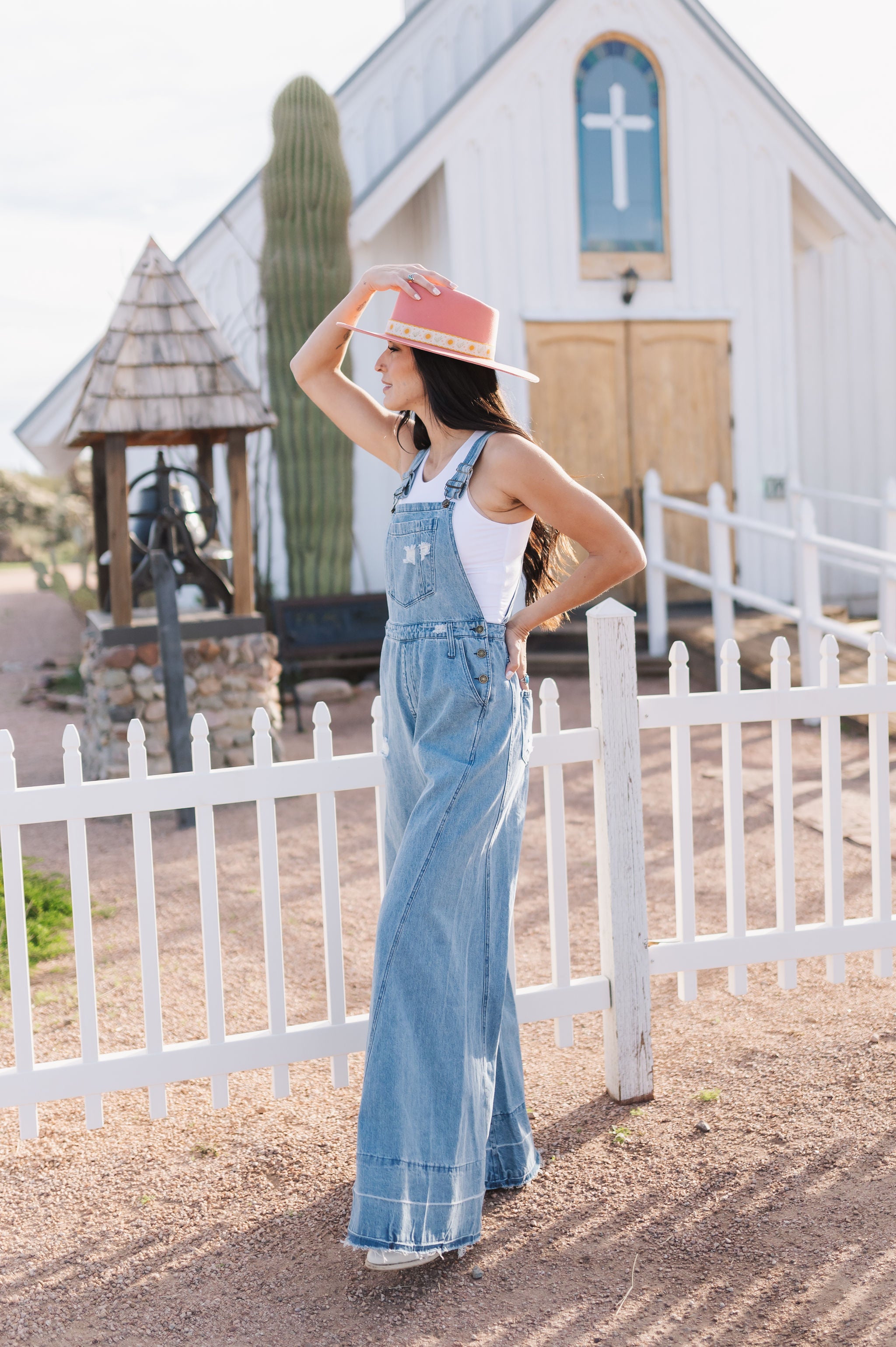 Side angled view of Clementine Wide Leg Distressed Denim Overalls with raw hem and front and side pockets.