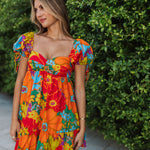 Front view of Paradise Blossom Mini Dress with allover bright tropical print, puff sleeves with ties, bustier top, and ruffle hem. 