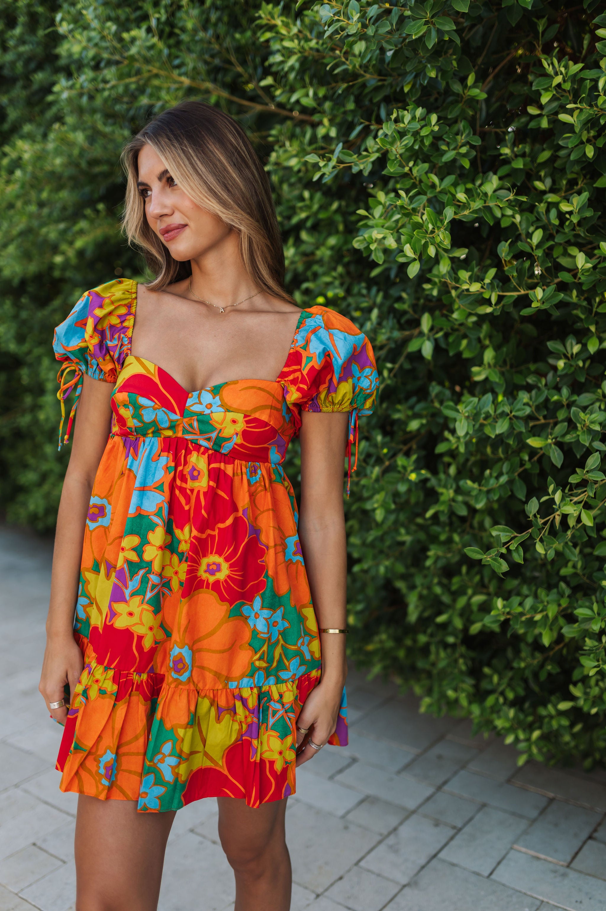 Front view of Paradise Blossom Mini Dress with allover bright tropical print, puff sleeves with ties, bustier top, and ruffle hem. 