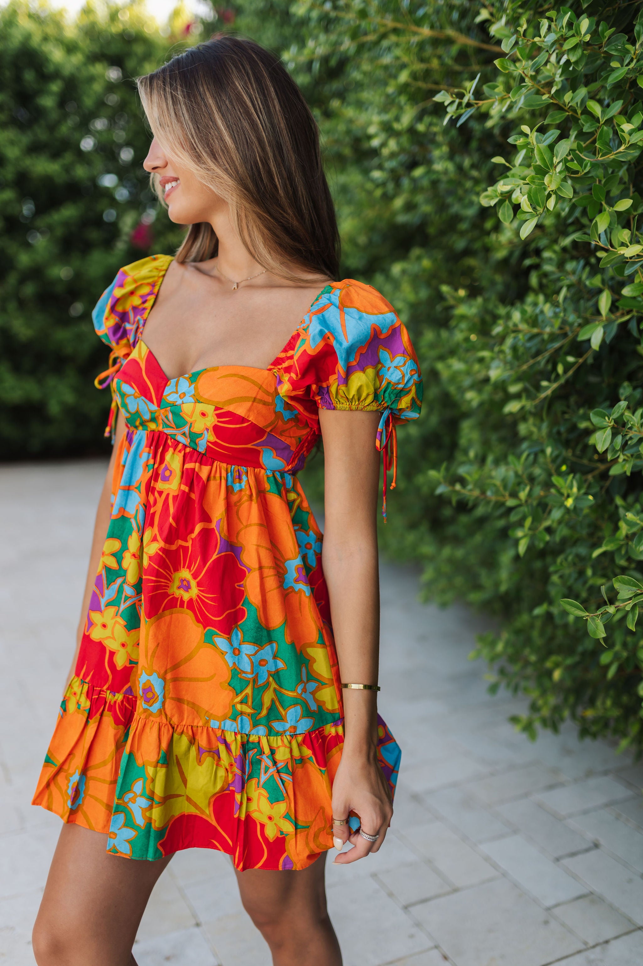 Front view of Paradise Blossom Mini Dress with allover bright tropical print, puff sleeves with ties, bustier top, and ruffle hem. 