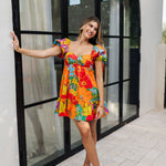 Front view of Paradise Blossom Mini Dress with allover bright tropical print, puff sleeves with ties, bustier top, and ruffle hem. 