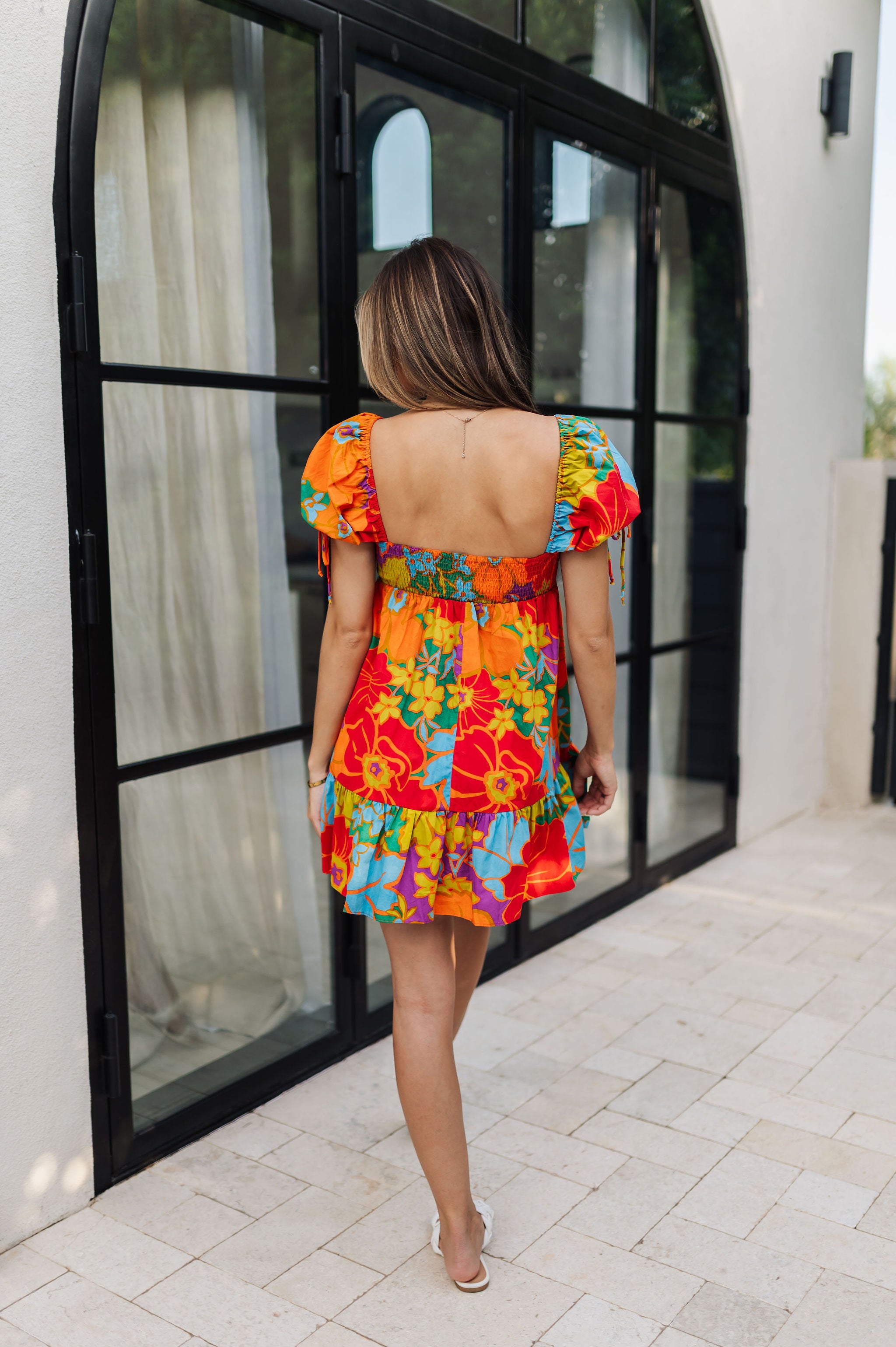 Rear view of Paradise Blossom Mini Dress with allover bright tropical print, puff sleeves with ties, smocked back, and ruffle hem. 